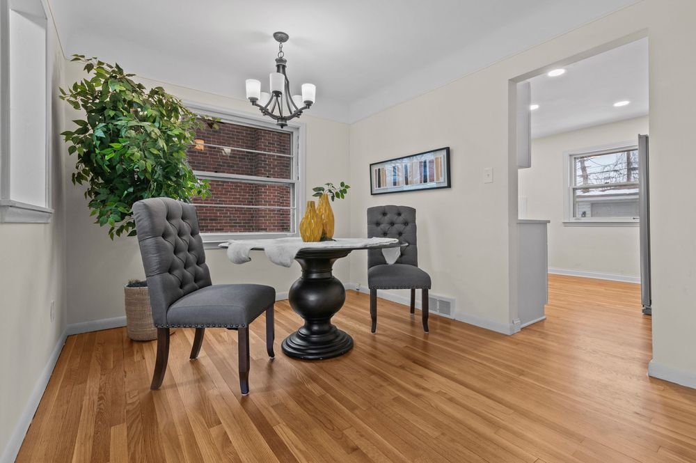 Elegant dining area with hardwood floors, tufted gray chairs, round pedestal table, chandelier, and large window with natural light.