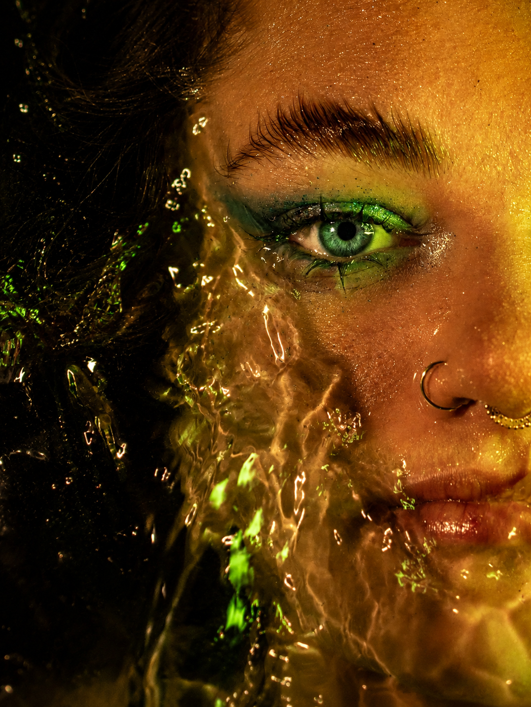 Close-up portrait of a woman with green eye makeup and nose ring, illuminated by golden water splashing across her face.