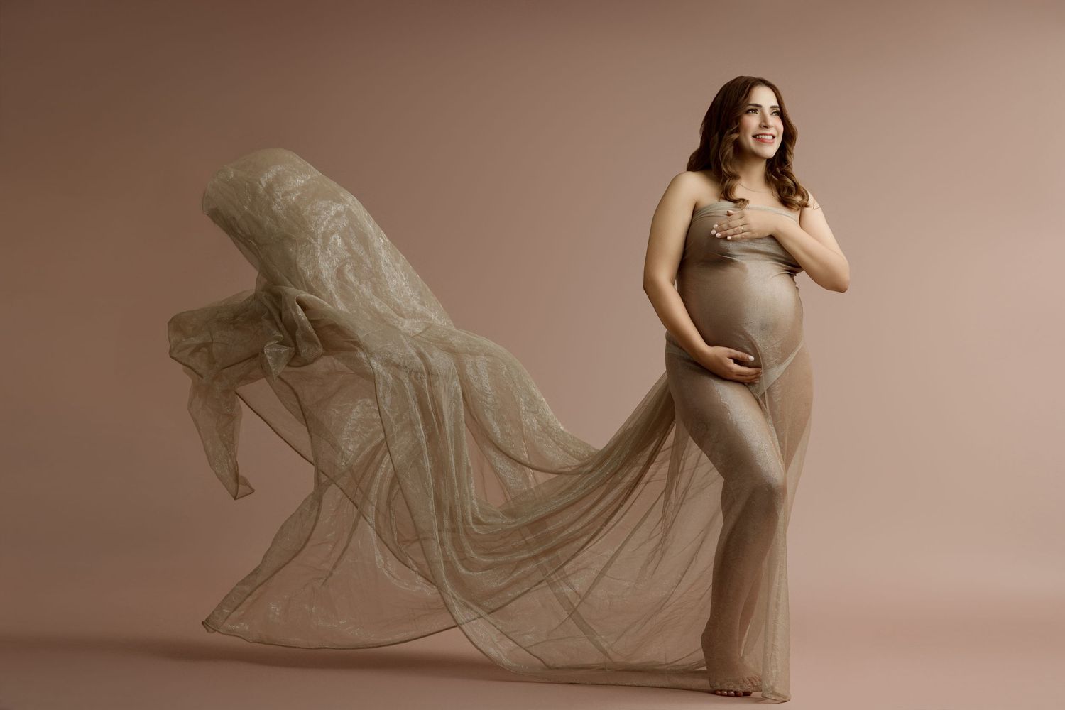 Expectant mother in a long gown posing against a warm neutral backdrop