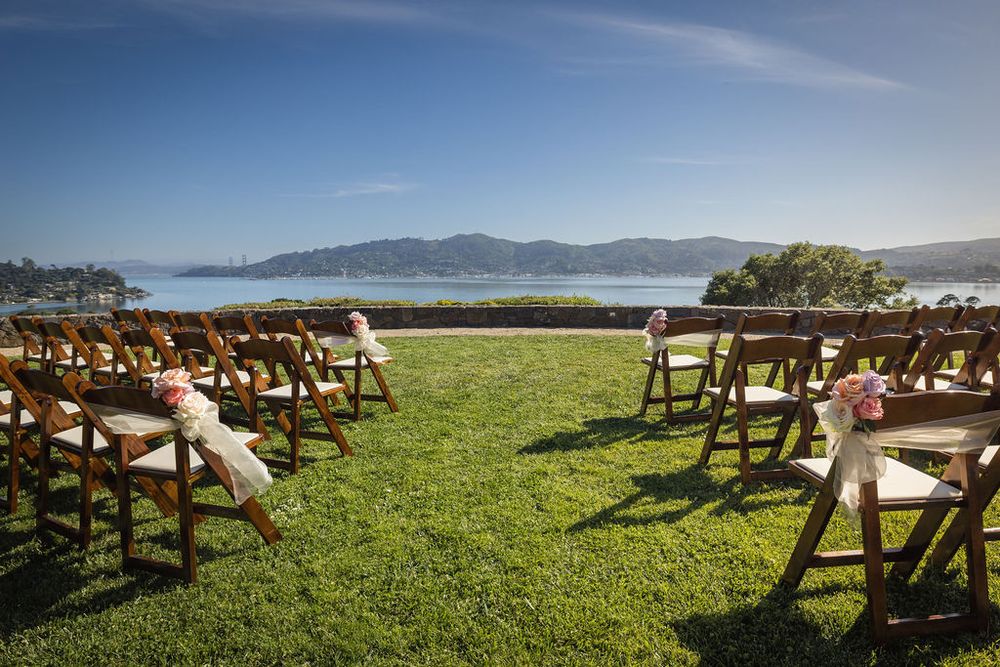Outdoor wedding ceremony setup with wooden chairs on green lawn overlooking a scenic bay and mountains under blue sky.
