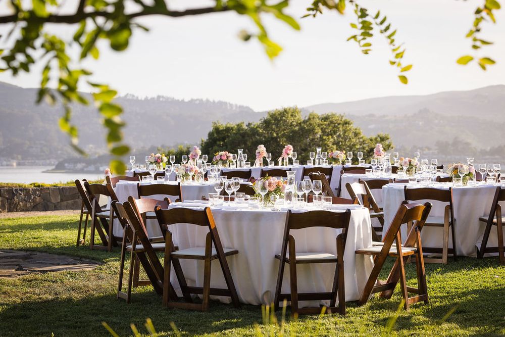 Elegant outdoor wedding reception with round tables, white linens, and wooden chairs set on a green lawn with scenic water views.
