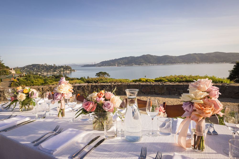 Elegant outdoor wedding table setting with pink roses and white linens overlooking a scenic bay and rolling hills.