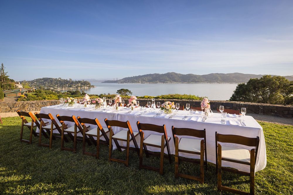 Elegant outdoor long dining table with white tablecloth set overlooking scenic bay and mountains under clear blue sky.