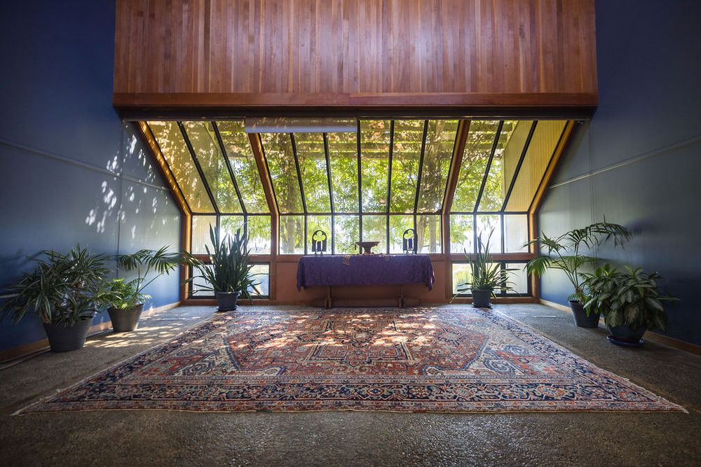 Elegant mid-century modern lobby with floor-to-ceiling windows, lush trees, ornate Persian rug, and a reception desk centerpiece.
