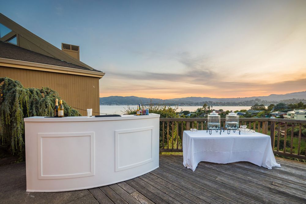 Elegant outdoor bar and catering setup on wooden deck overlooking scenic bay at golden sunset.