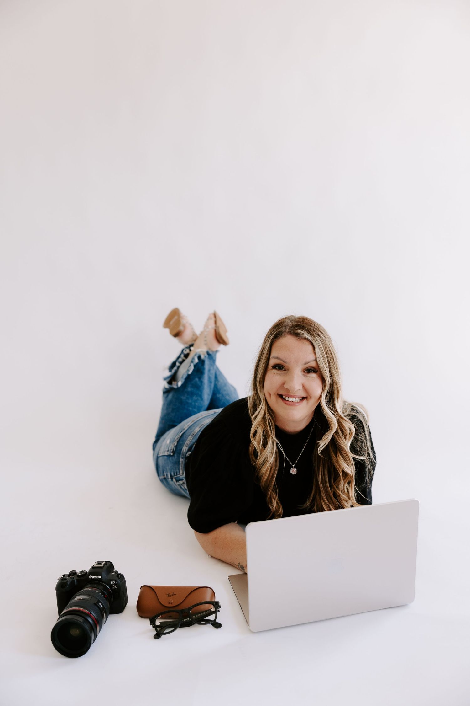 Smiling woman lying on white floor with laptop, DSLR camera, glasses, and brown wallet nearby.