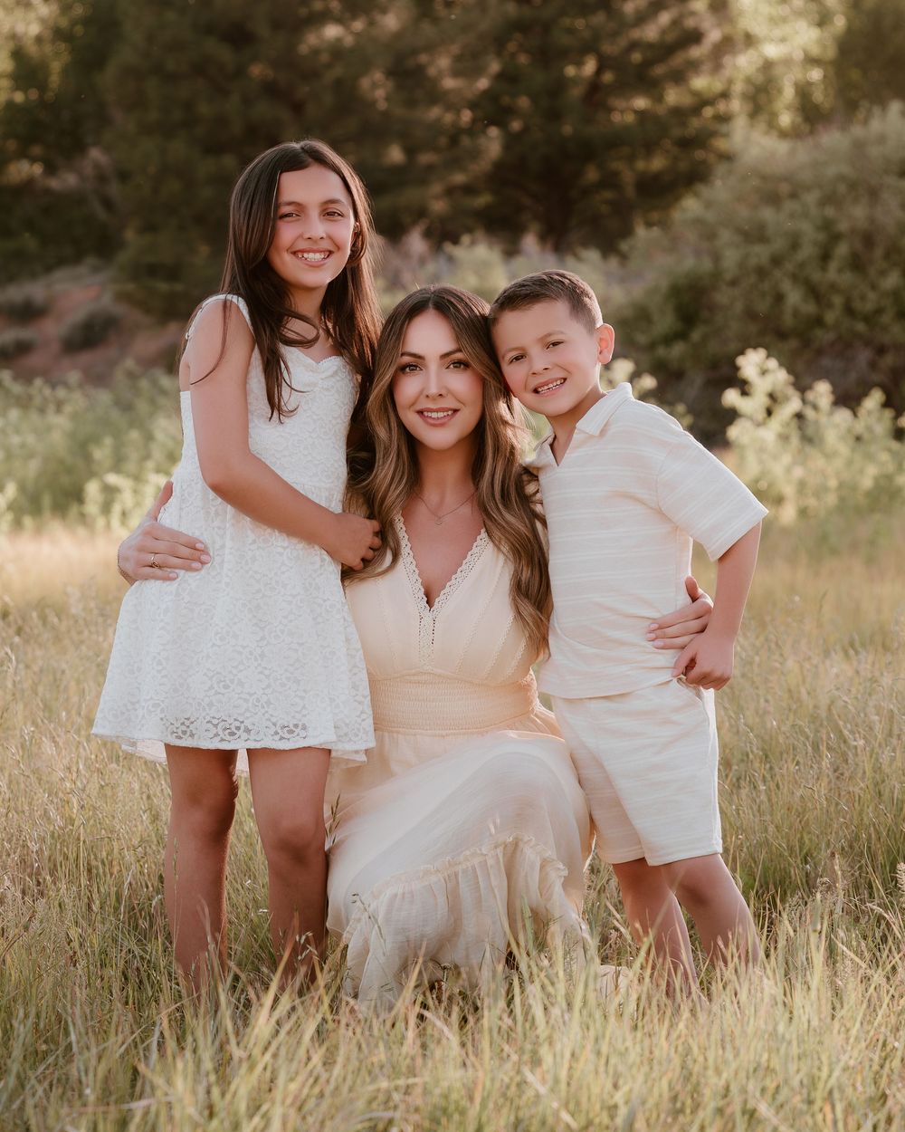 A woman kneels in a golden field with two children beside her, all wearing white and cream outfits, smiling warmly.