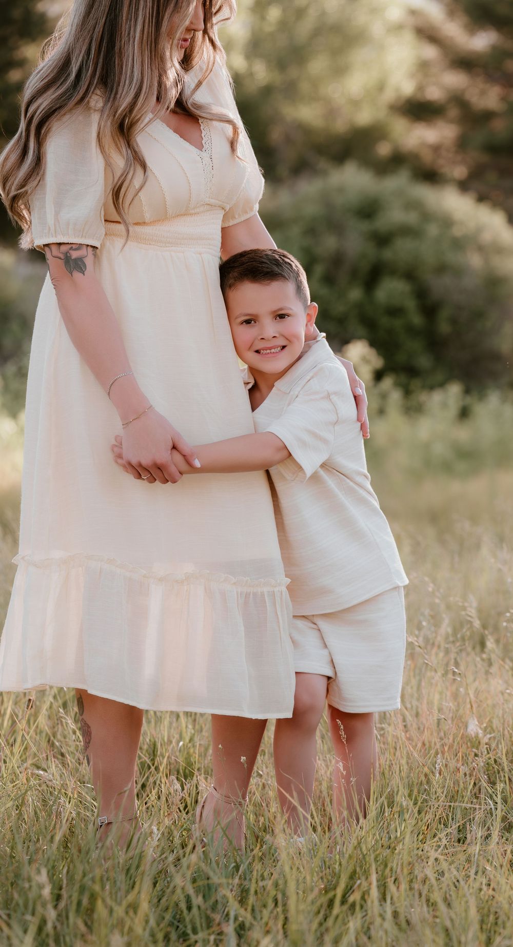 A young boy hugs his pregnant mother in a sunny field, both wearing matching cream outfits.