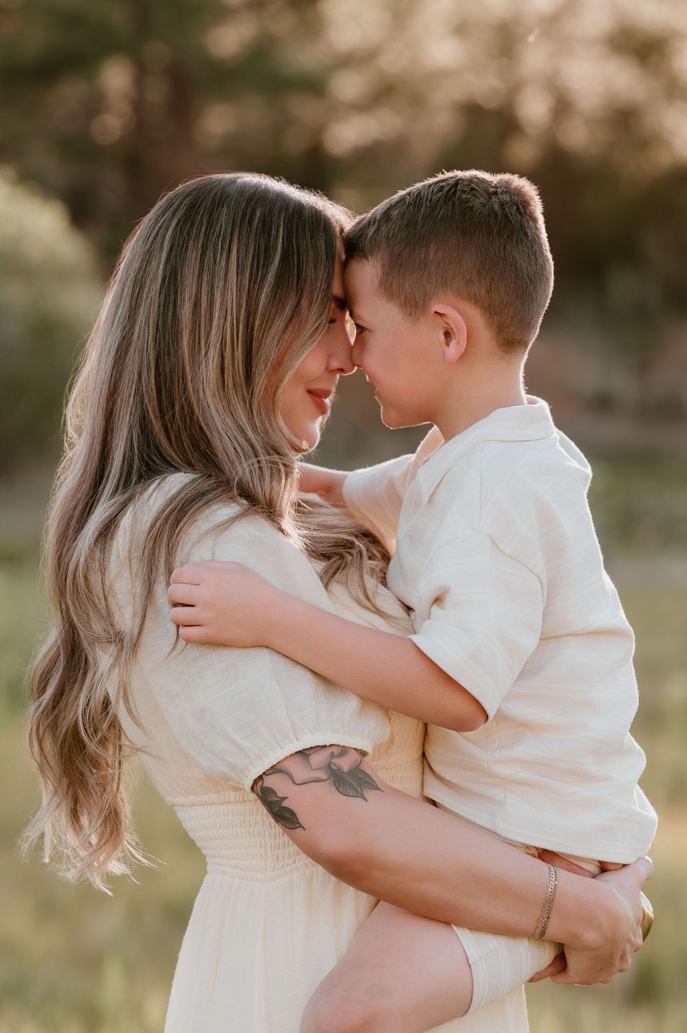 A mother in a white dress holds her young son close, touching foreheads in a tender outdoor embrace at golden hour.