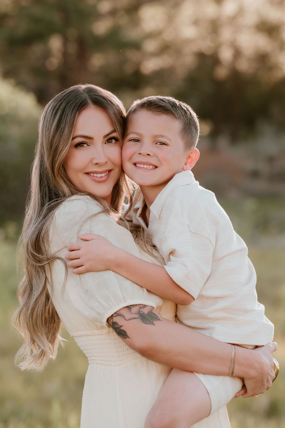 A smiling woman with long hair and tattoos holds a happy young boy outdoors in warm golden light.