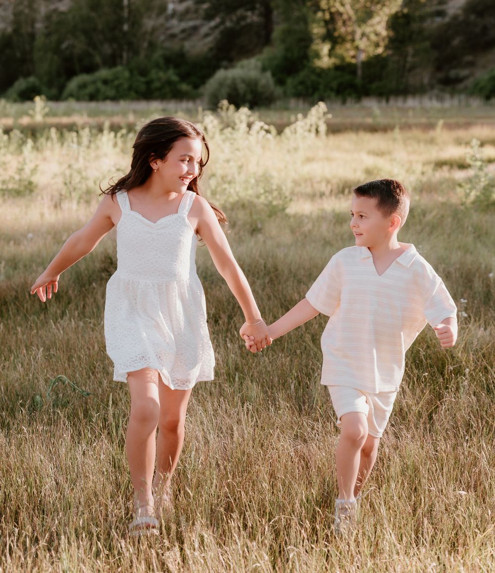 Two children in white outfits joyfully run hand-in-hand through a sunny golden meadow.