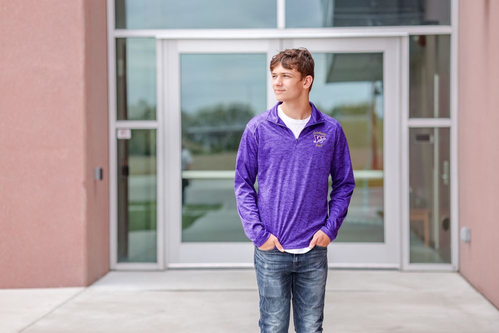 Senior in purple pullover walks with his hands in pockets in front of school doors