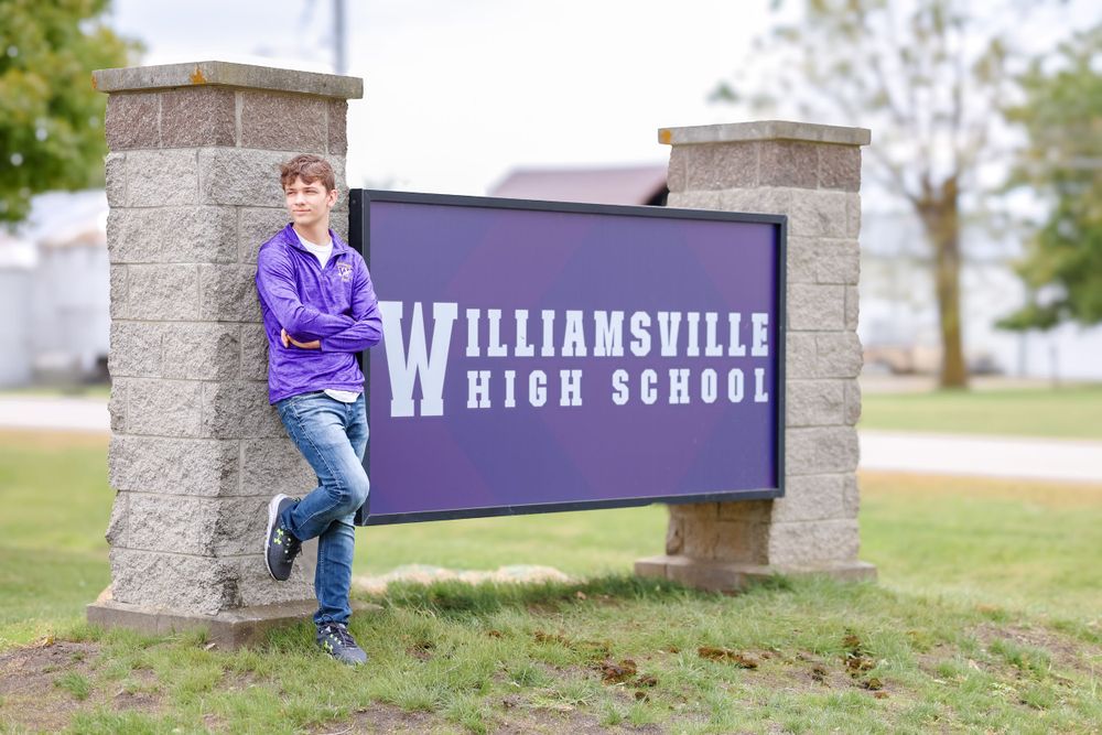 Senior in purple pullover stands against school sign with arms crossed