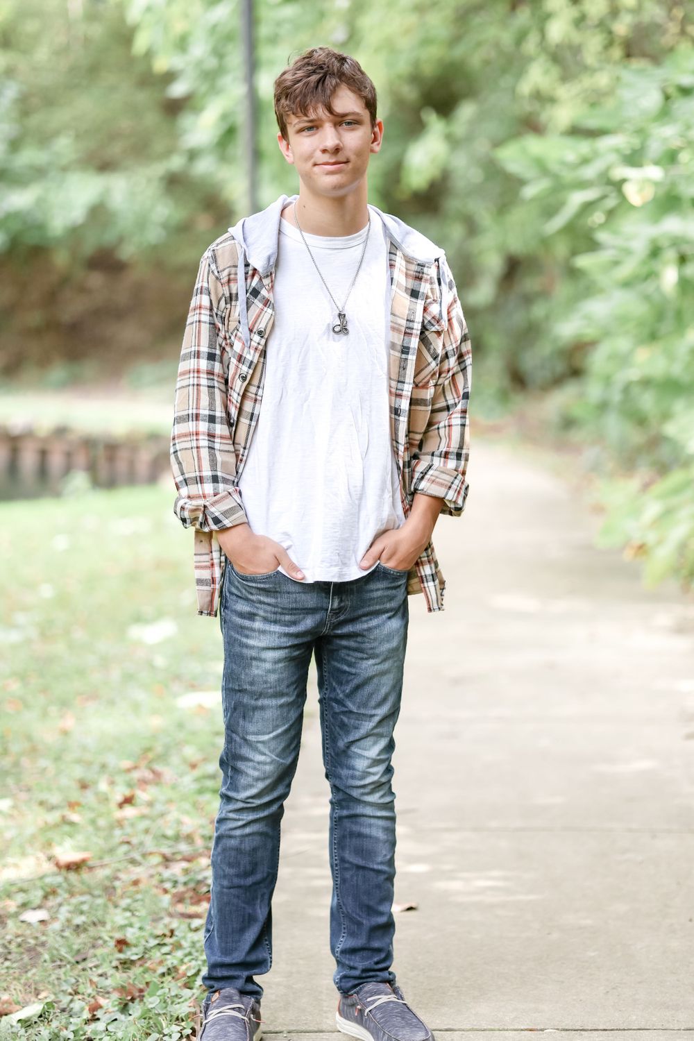 Senior in white shirt and tan plaid jacket stands with his hands in pockets while standing on a sidewalk.