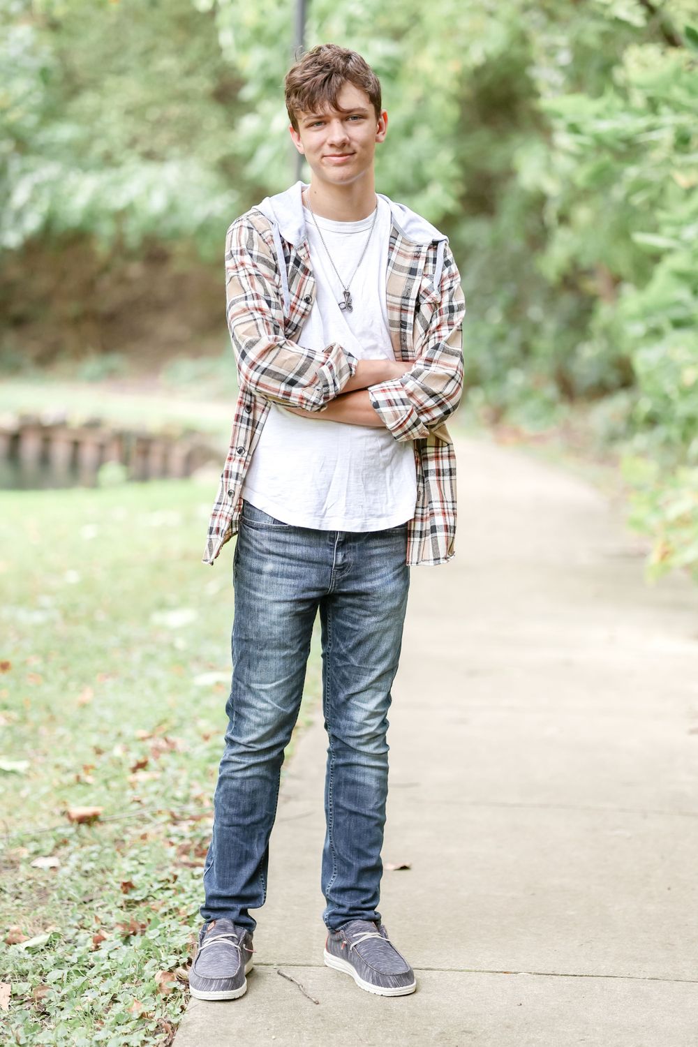 Senior in white shirt and tan plaid jacket stands with arms crossed on a sidewalk outdoors