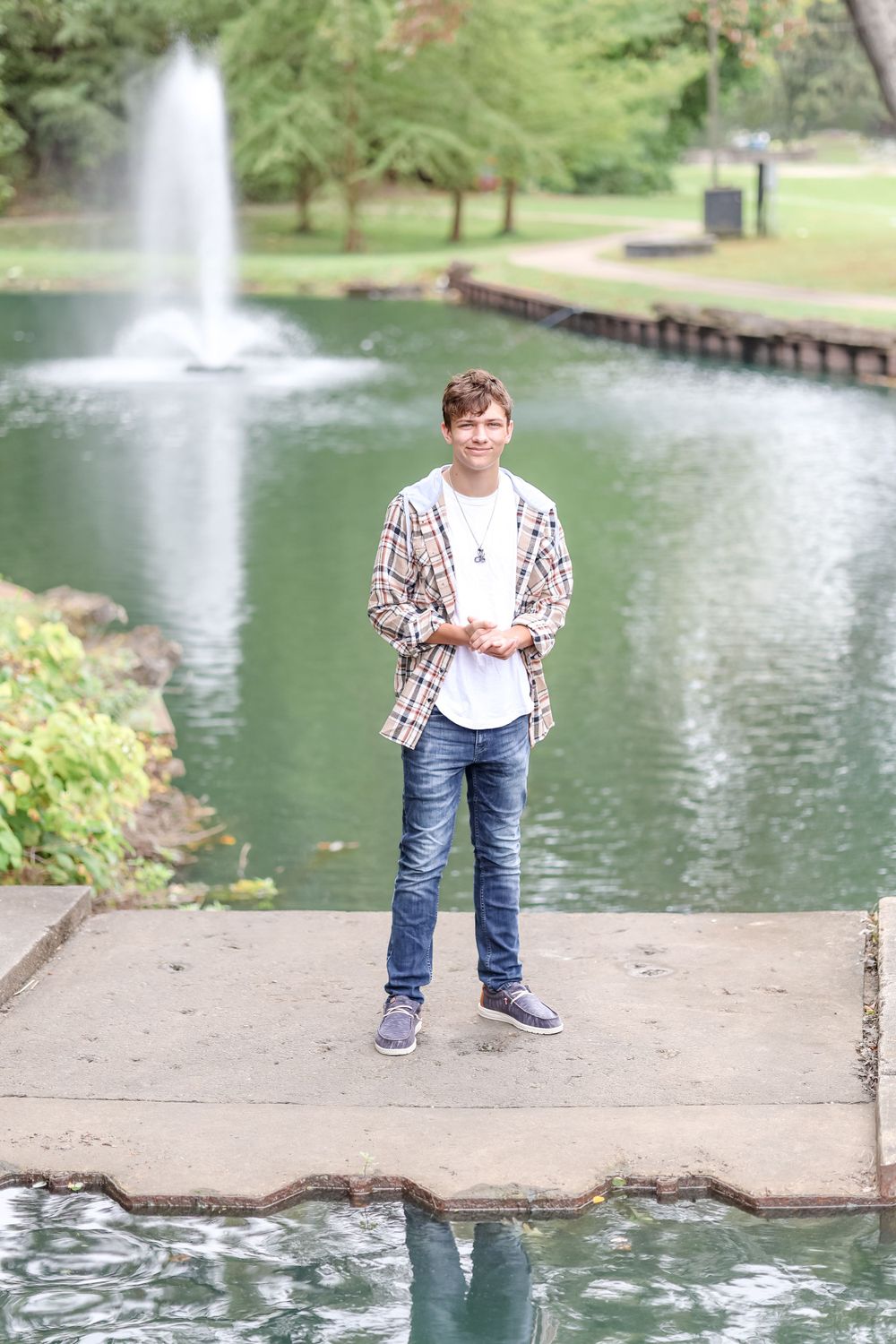 Senior in white shirt and tan plaid jacket stands on concrete pathway in front of pond with a fountain