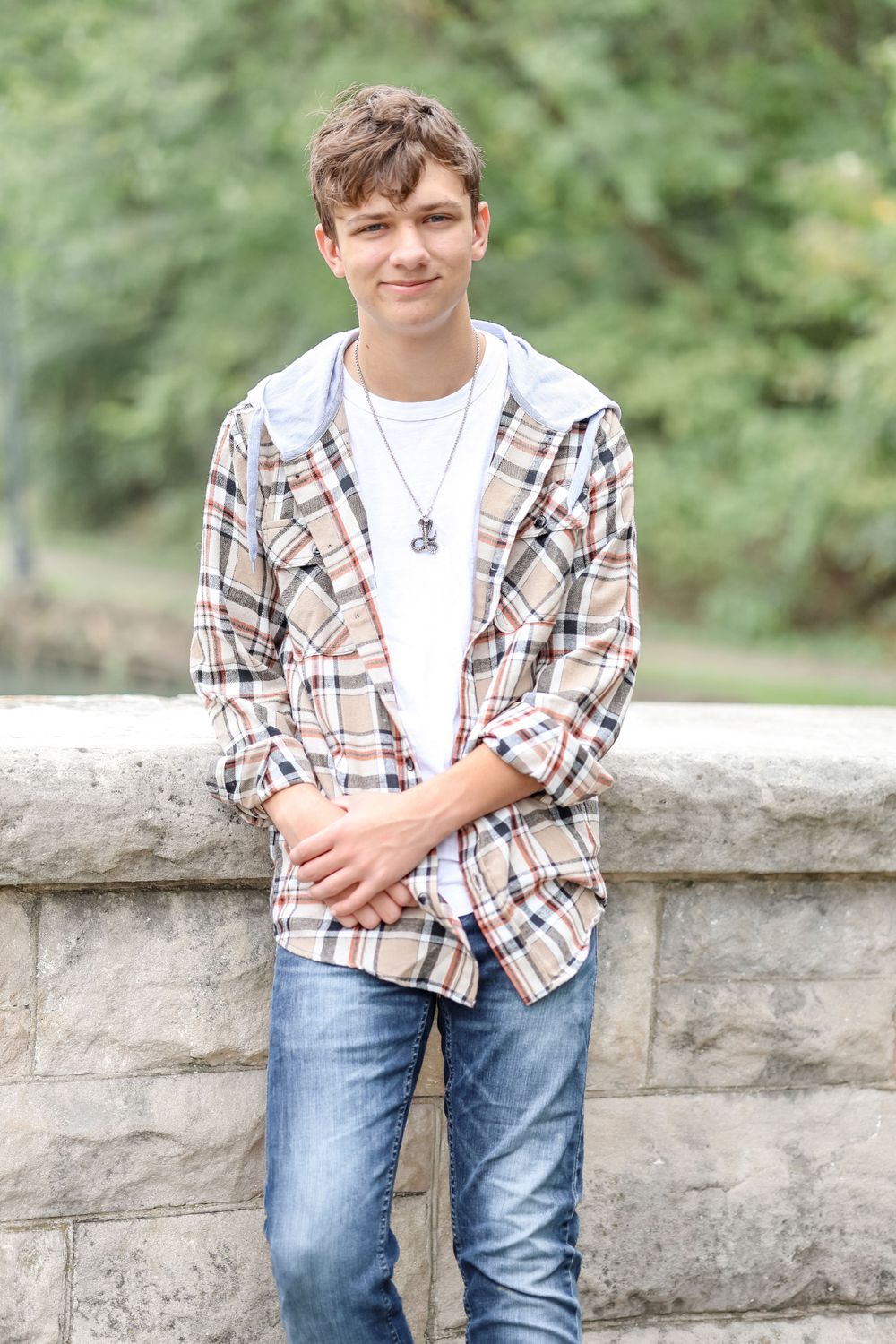 Senior in white shirt and tan plaid jacket stands and leans against a stone bridge