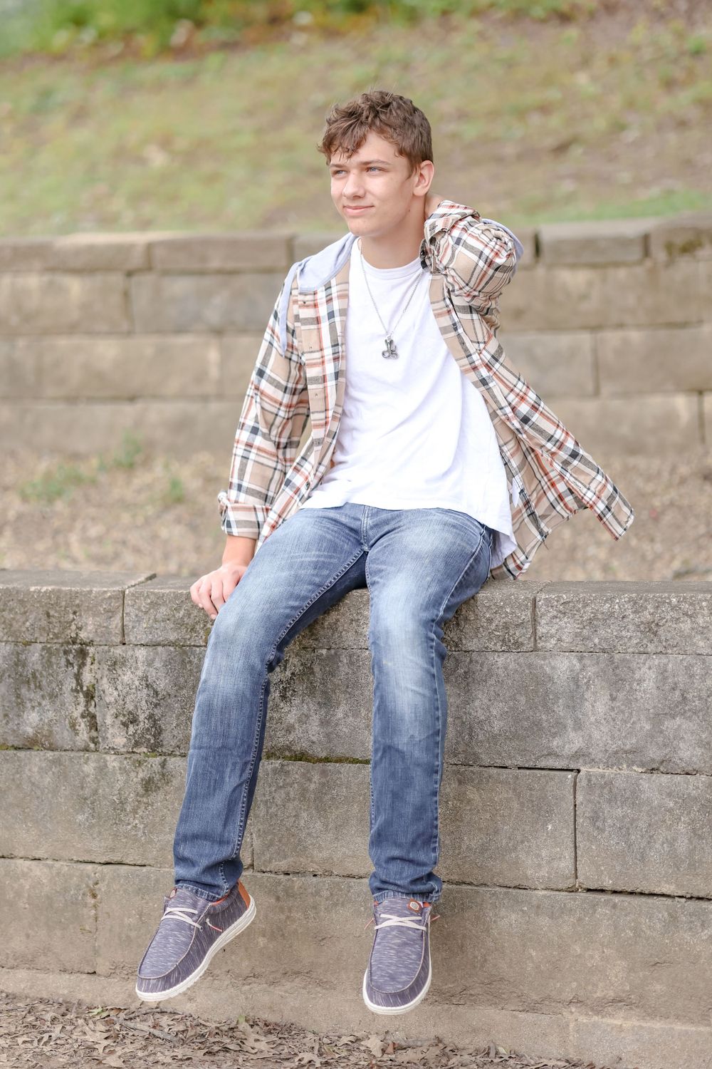 Senior in white shirt and tan plaid jacket sits on ledge of brick wall with one hand behind head