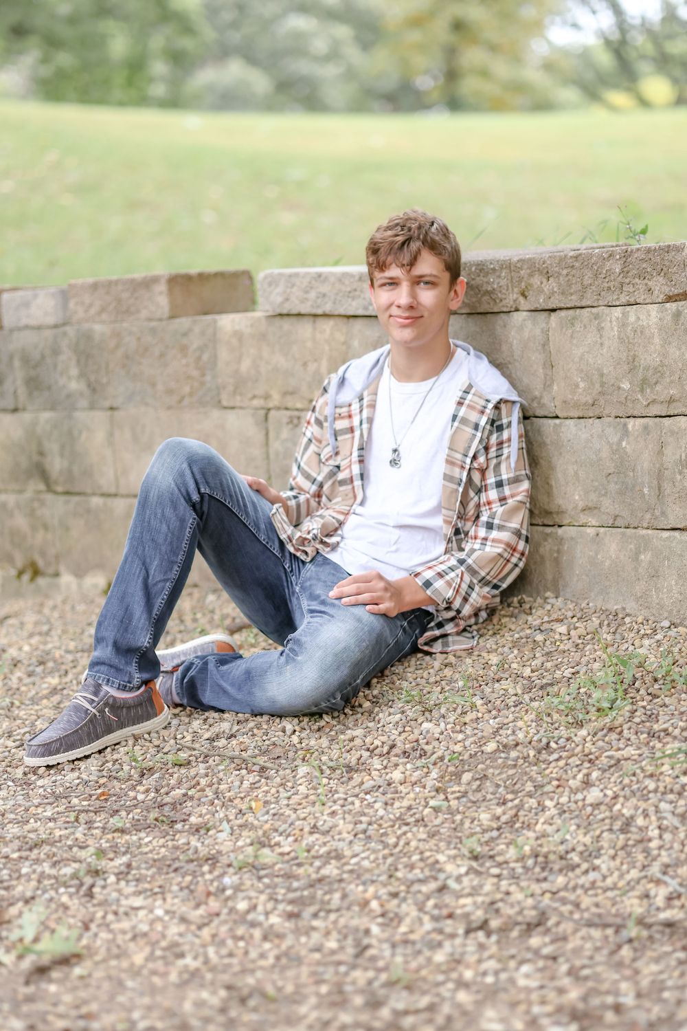 Senior in white shirt and tan plaid jacket sits against a small brick wall