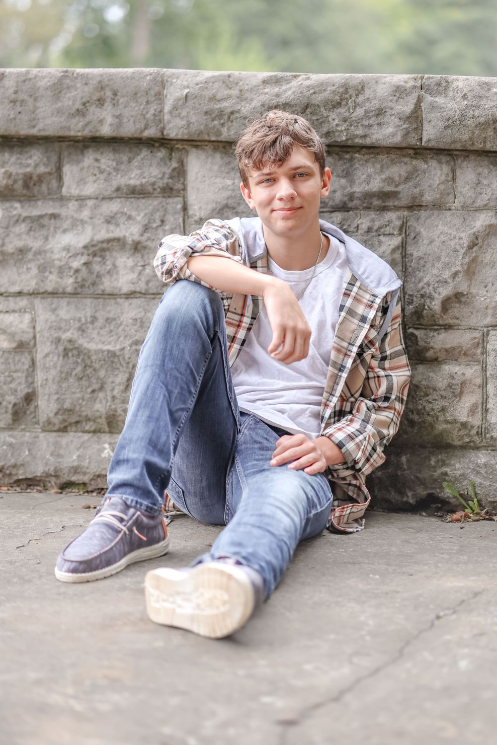 Senior in white shirt and tan plaid jacket sits on ground leaned against a stone wall