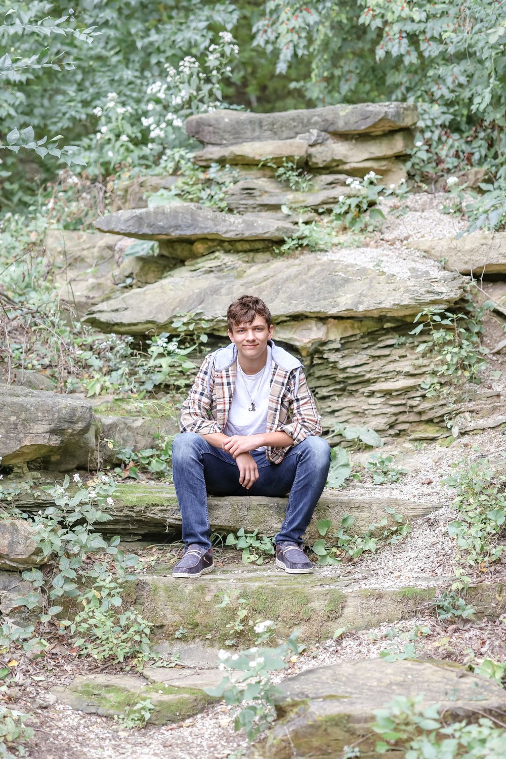 Senior in white shirt and plaid jacket sits on large stone rocks
