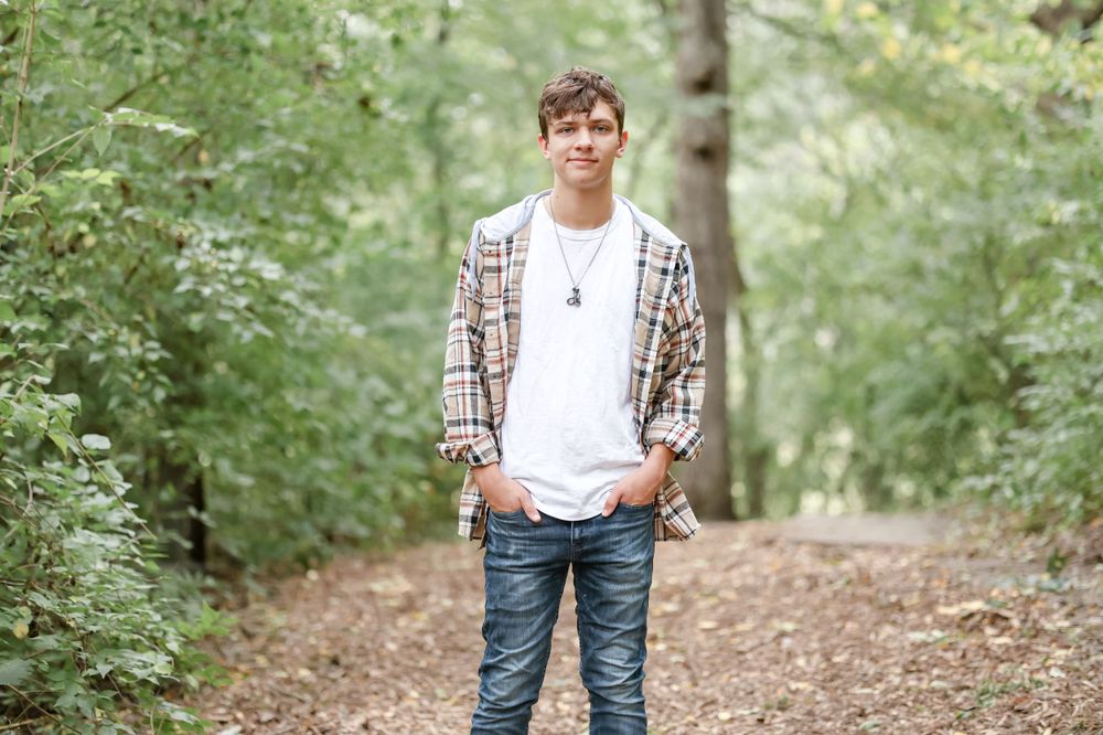Senior in white shirt and plaid jacket stands with hands in his pocket on a trail