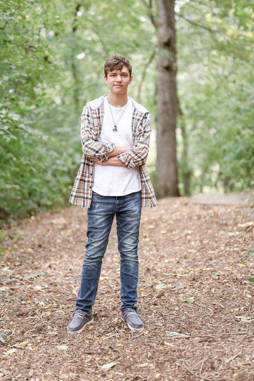 Senior with white shirt and plaid jacket stands and crosses arms on trail surrounded by greenery