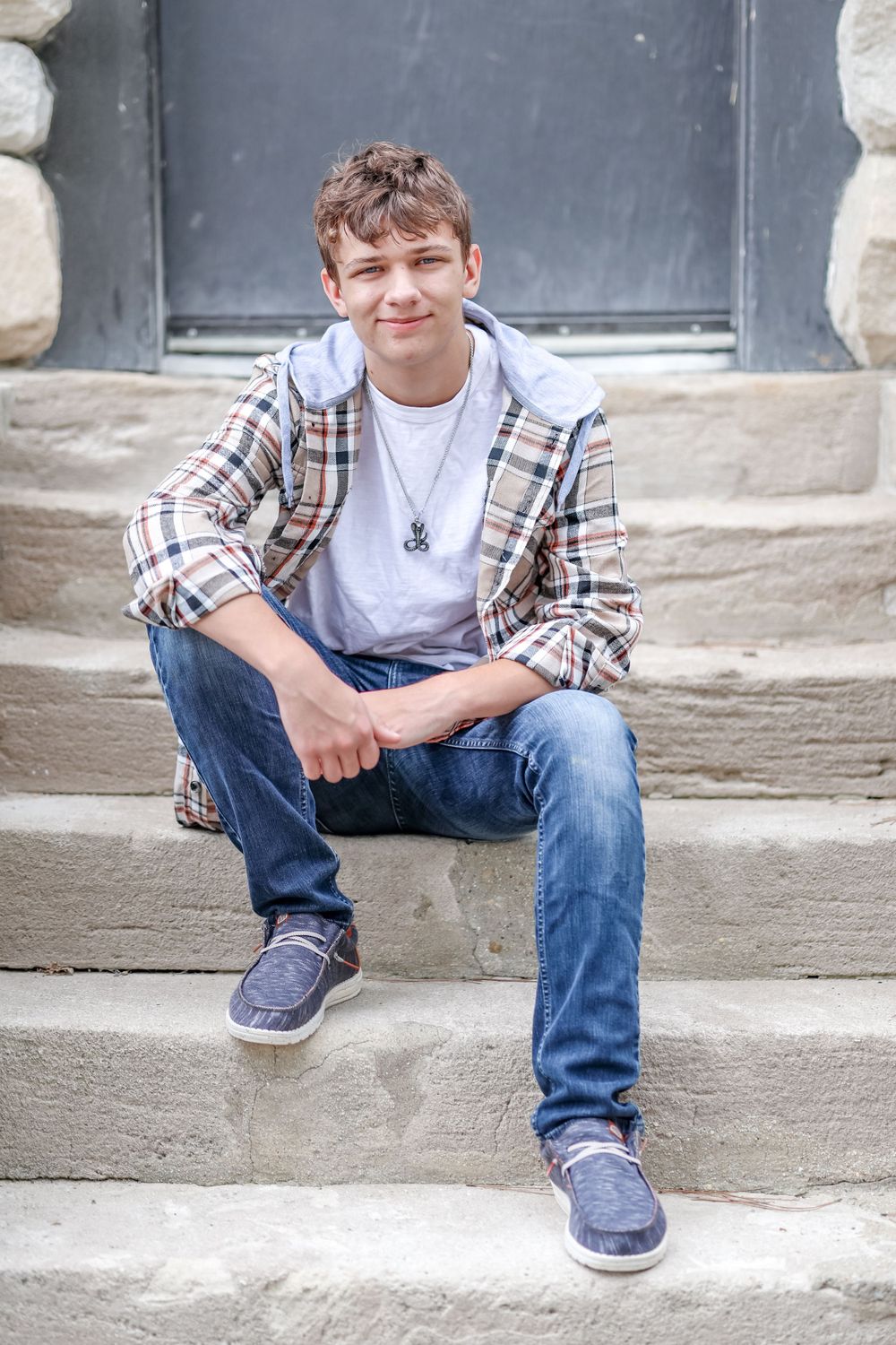 Senior in white shirt and plaid pullover sit on steps in front of a black door