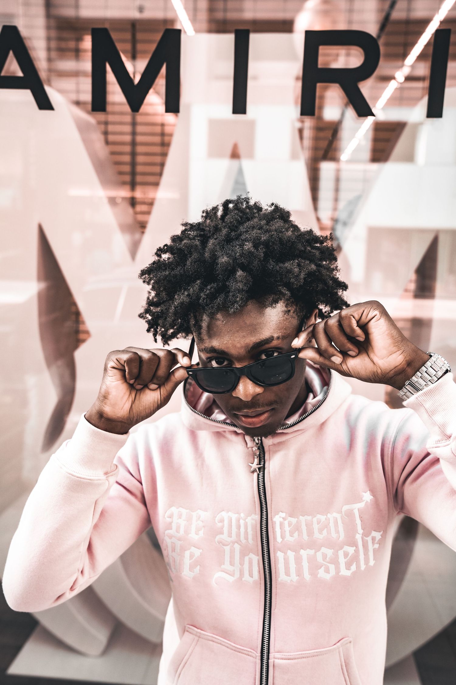 Fashion portrait with pink hoodie and sunglasses against AMTR lettering background.