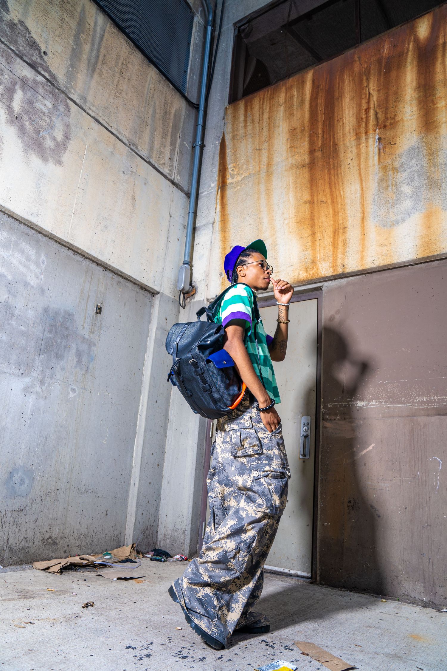 Person in striped polo shirt and camo pants posing against industrial wall.