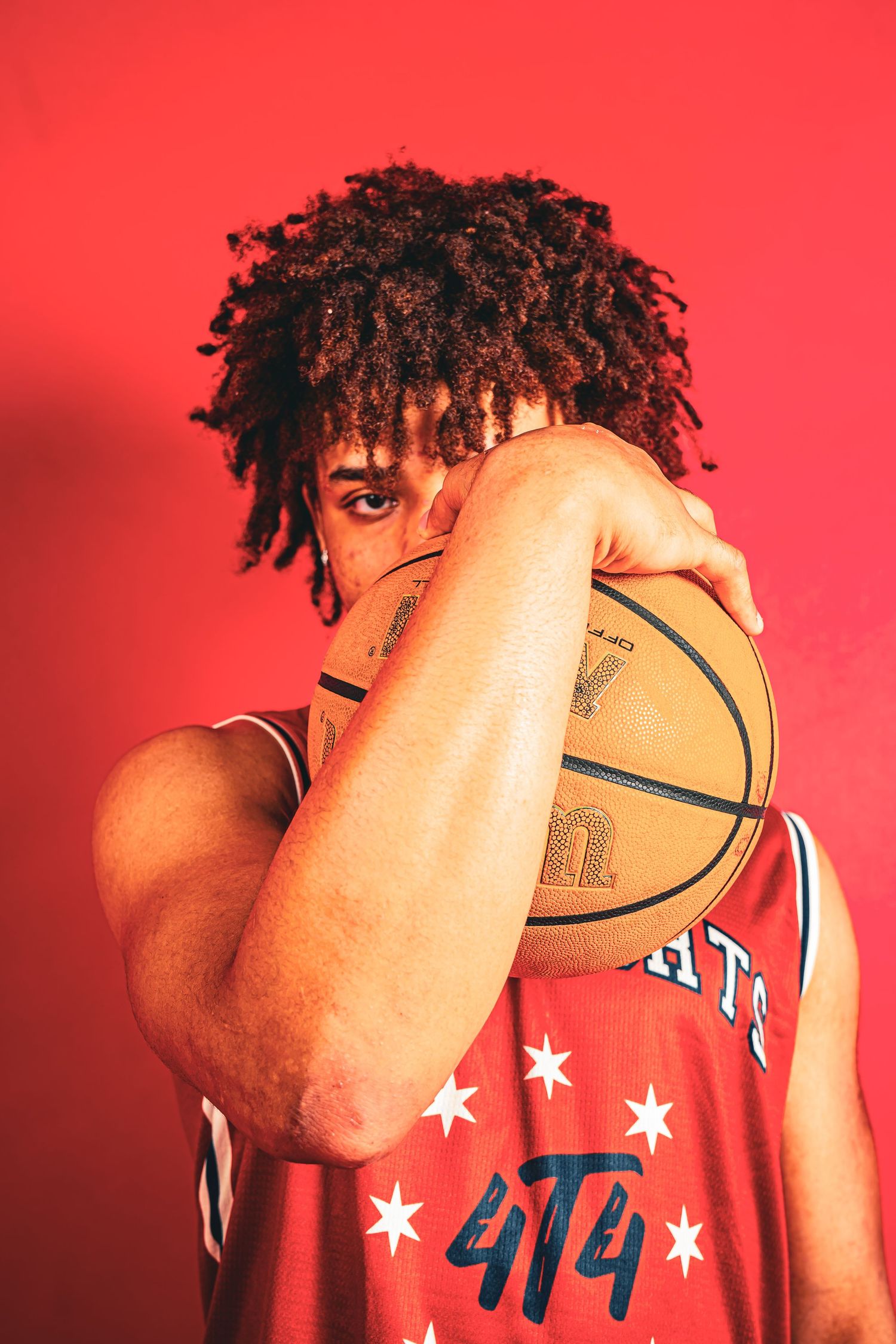 Basketball player with curly hair poses with ball wearing red jersey against red background.
