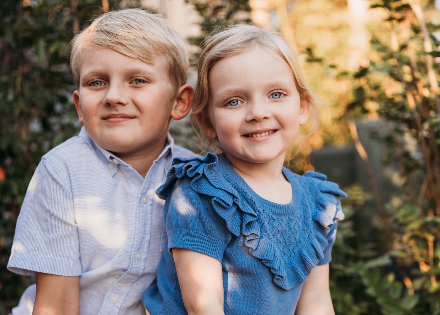 Two children in casual clothing pose outdoors with golden sunlight filtering through trees.