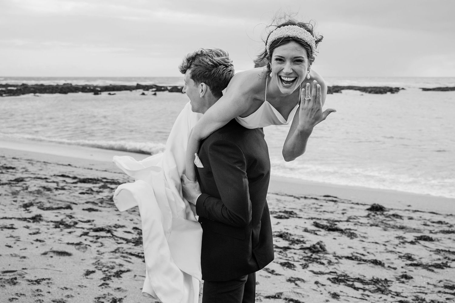 wedding couple on devon beach having fun whilst groom lifts bride and carries her whilst she shows off her wedding ring