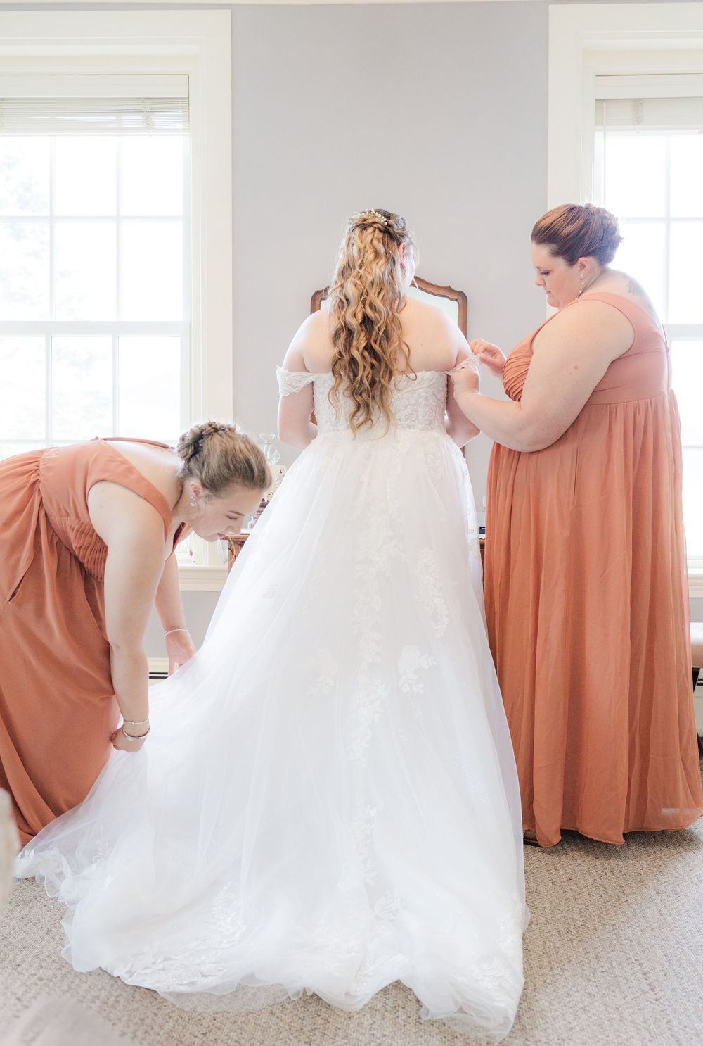 Bridesmaids in coral dresses assisting with wedding gown preparations.