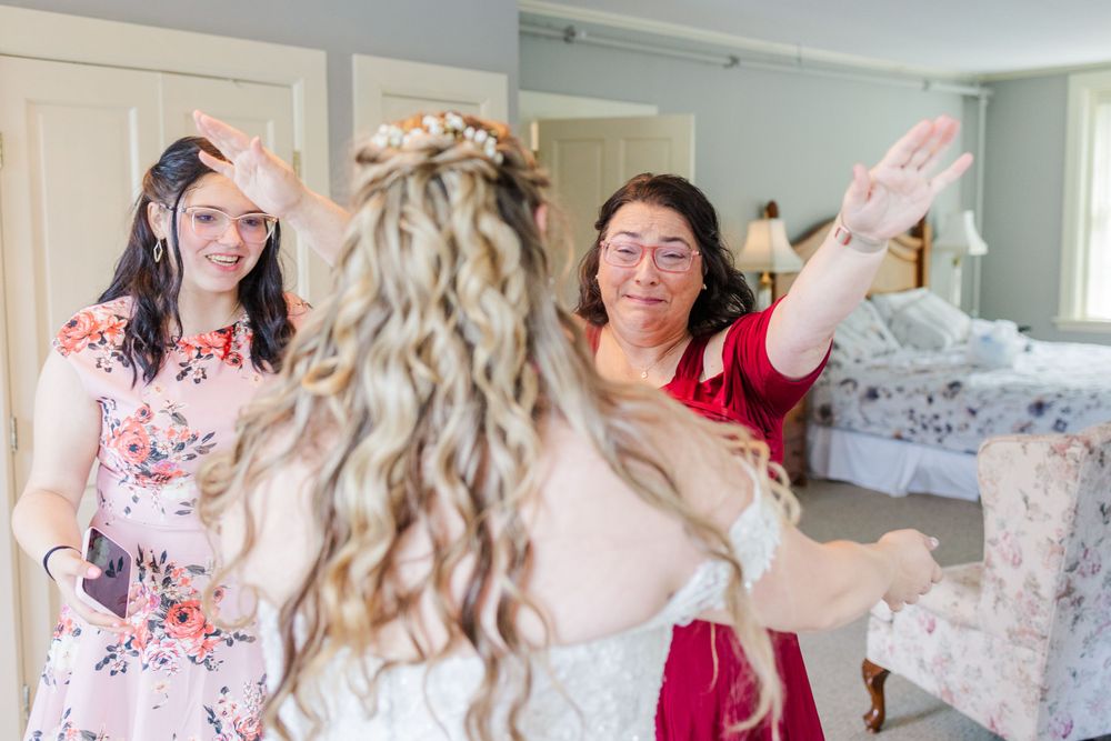 Friends laugh and celebrate as they help prepare a bride in her white floral wedding gown.