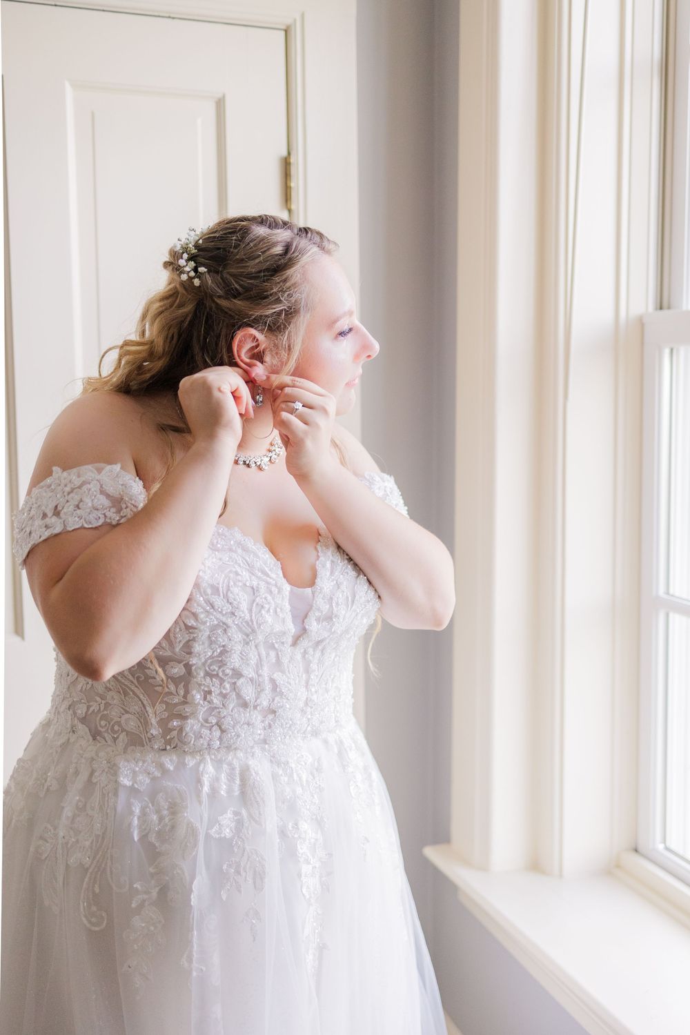 Bride in lace wedding dress puts on earrings while standing by window in natural light.