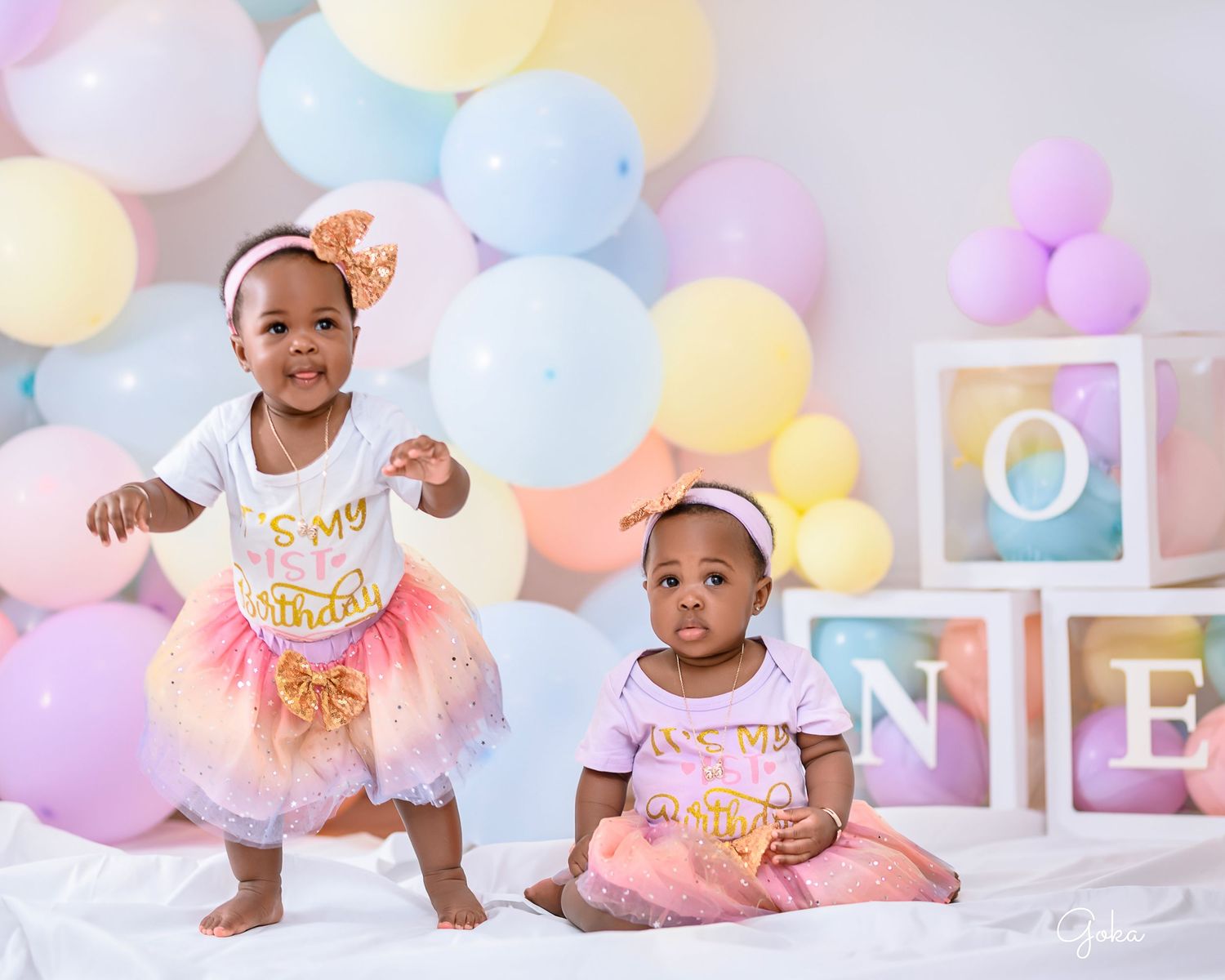 Two toddlers in tutus pose for a birthday photo with pastel balloons and letter blocks.