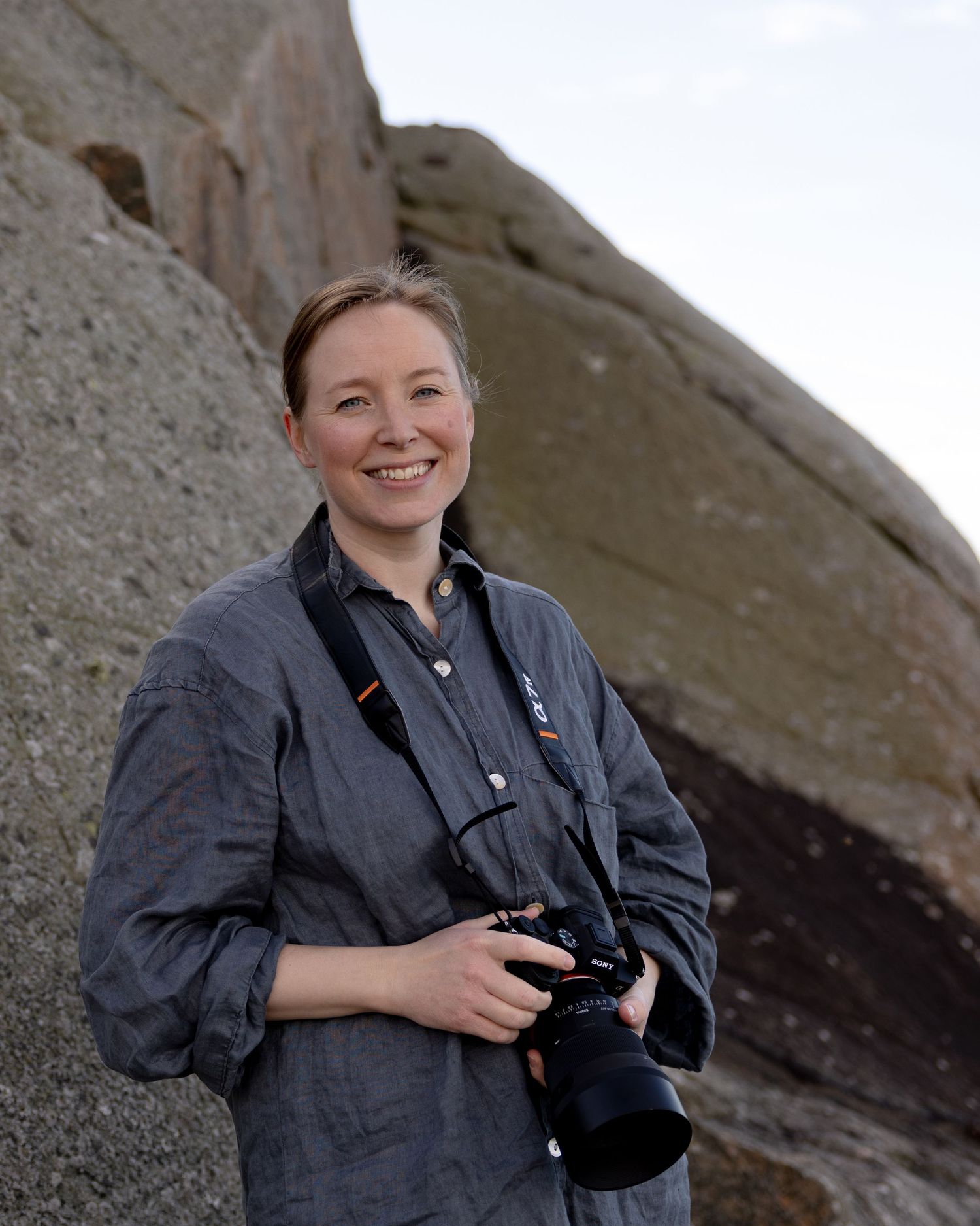 Fotografen Emma Lönn håller i sin kamera och tittar in i kameran och ler. I bakgrunden syns gråa klippor.