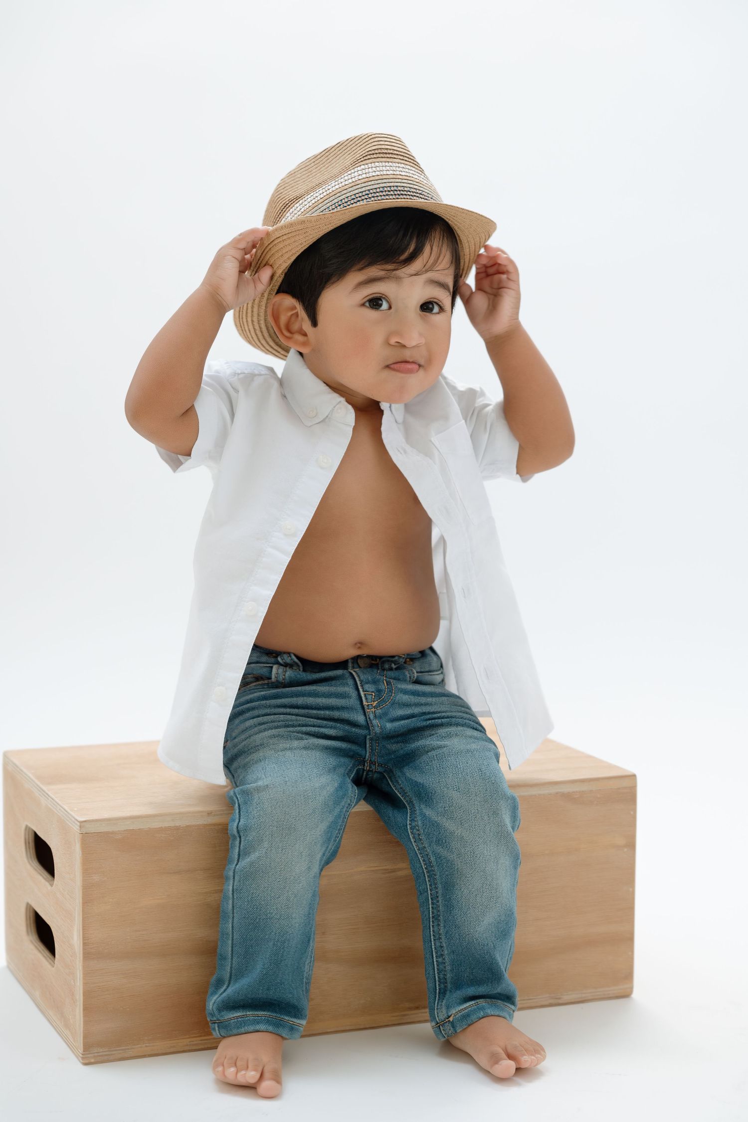 Small child wearing a straw hat, white shirt and jeans sits barefoot on wooden box against white background.