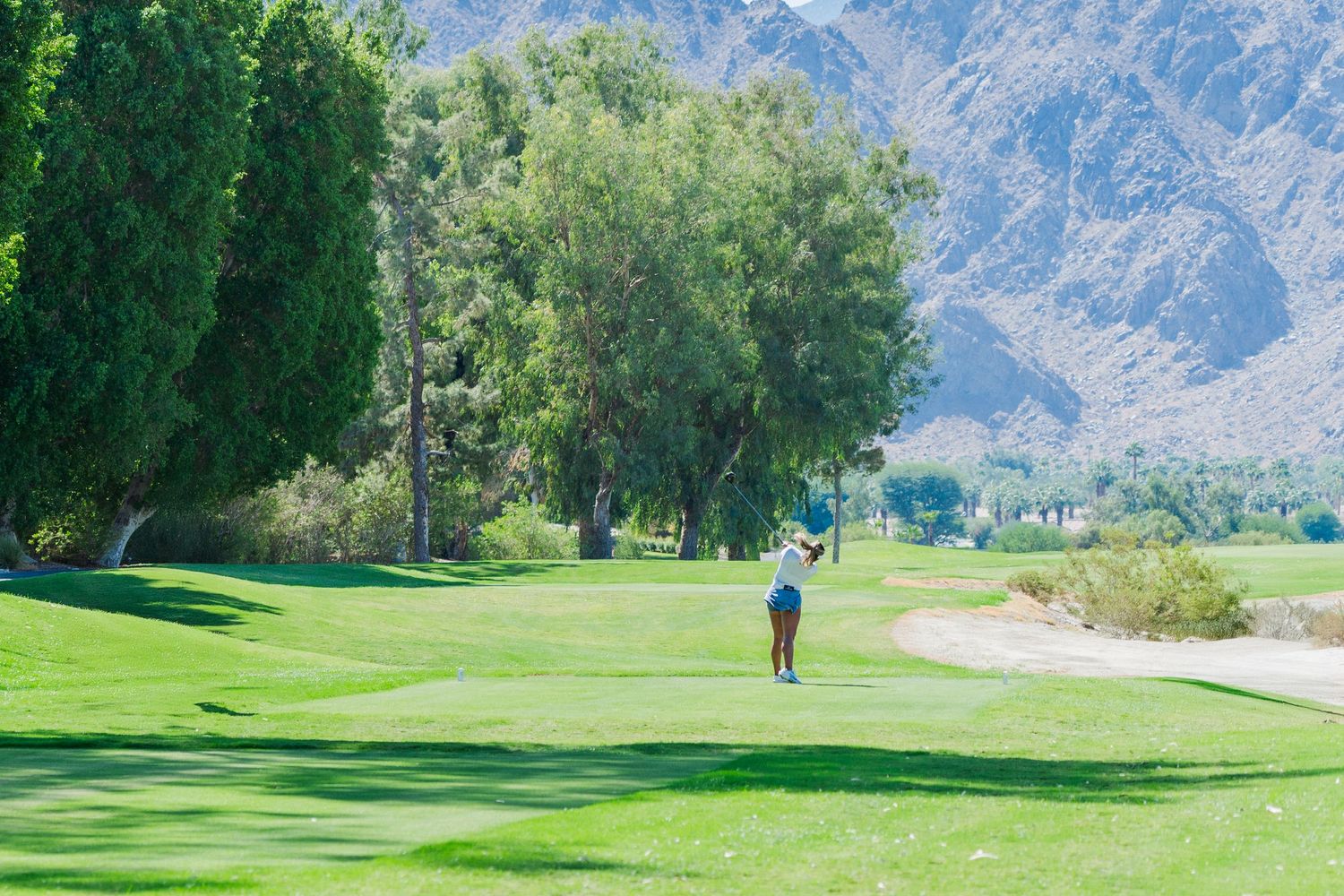 Golfer swings on lush green course with majestic mountains and trees in the background.