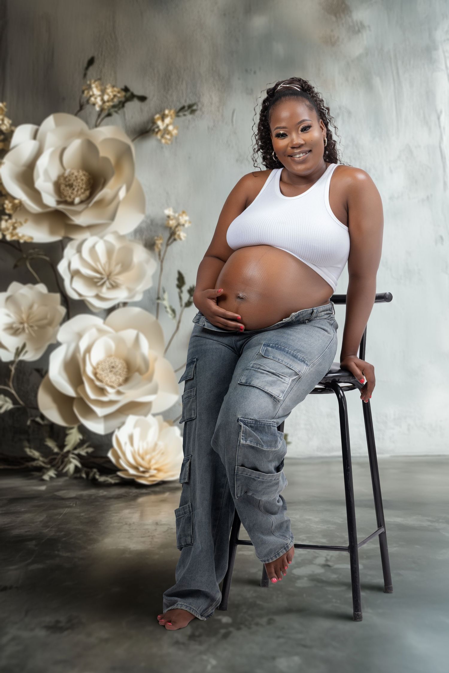 Maternity photoshoot with white tank top and cargo pants against floral wall backdrop.