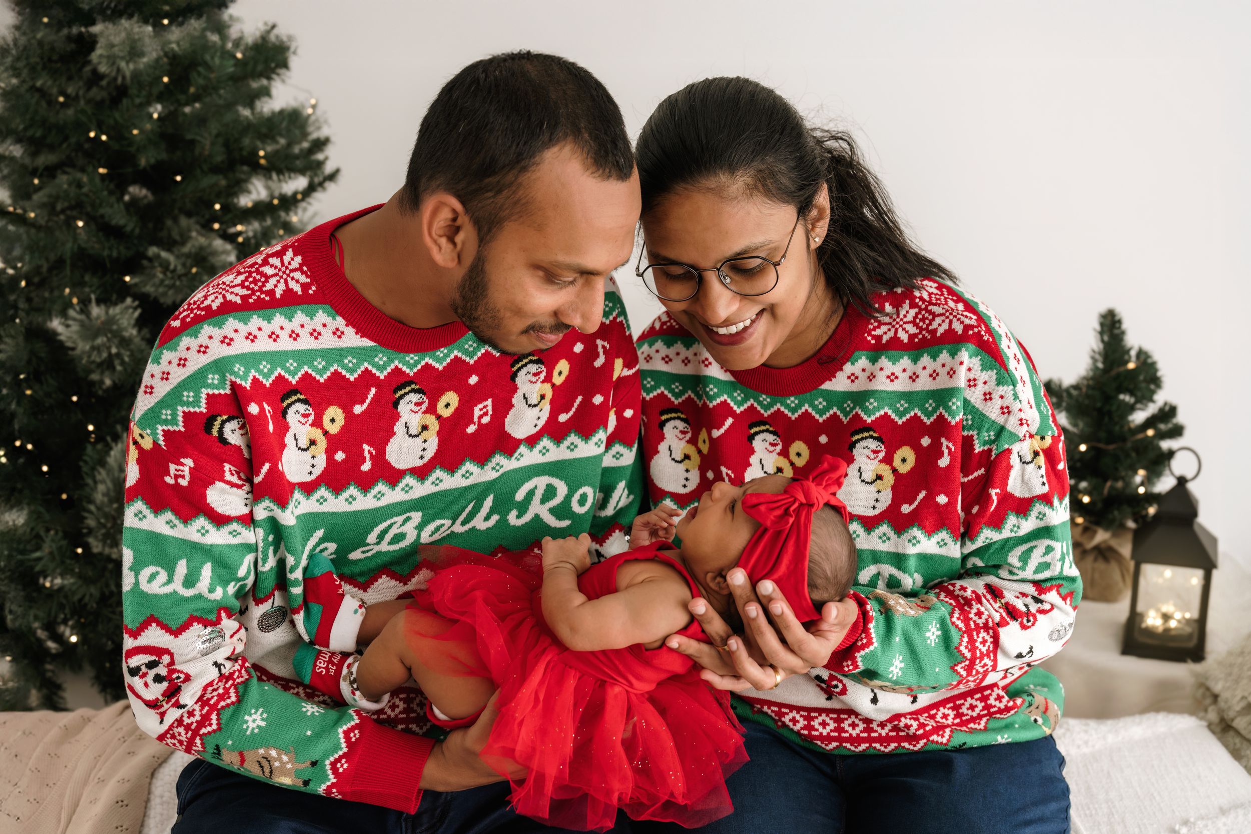 A couple wearing matching holiday sweaters cuddles together on a couch near a Christmas tree.