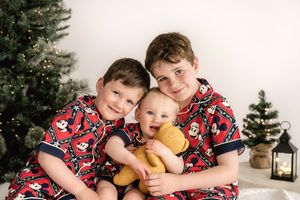 Three children in matching red holiday pajamas sitting together by a Christmas tree with decorative lanterns nearby.