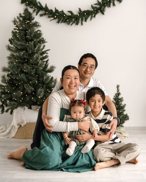 A family sits together on the floor in front of Christmas trees in a minimalist white studio setting.