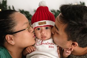 Parents kiss their baby wearing a red knit winter hat and white Christmas sweater during holiday photo session.
