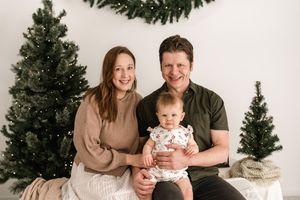 A family poses together in coordinating green and white outfits in front of a Christmas tree for holiday photos.