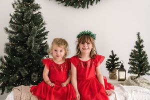 Two children in red dresses sit and pose together in a festive holiday setting with Christmas trees and decorations.