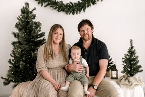 Family poses for holiday portraits in front of Christmas trees wearing coordinated neutral outfits.