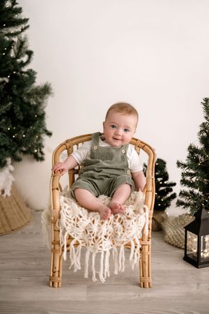 A baby sits in a rattan chair with macrame decor and Christmas trees in the background.
