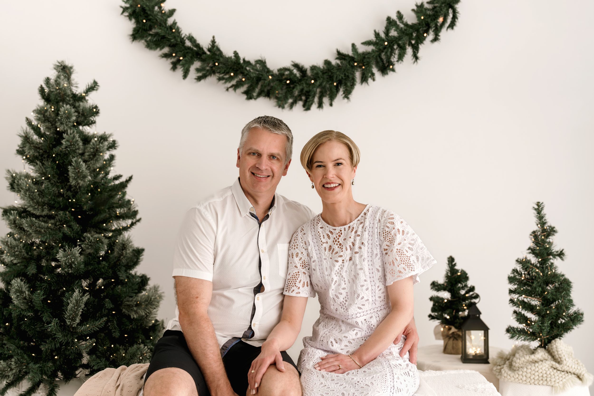 Couple in white outfits sits and poses together for holiday photos with Christmas trees and garland decorations.